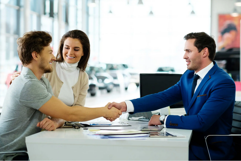 A couple is shaking hands with a salesperson in a car dealership office.