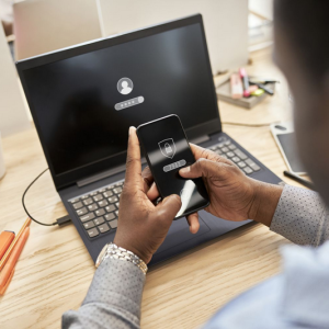 A person is entering a password on their phone in front of a laptop displaying a login screen.