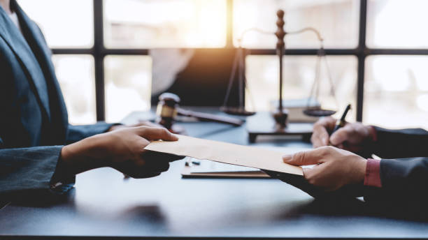 Two business professionals exchange documents in an office with a gavel and scales of justice in the background.