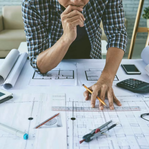 A person is studying architectural blueprints on a desk cluttered with drawing tools, a tablet, a calculator, and eyeg